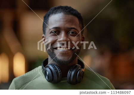 Front view portrait of African American young man with headphones smiling at camera blurred background copy space Front view portrait of African American young man with headphones smiling at camera blurred background copy space 113486097