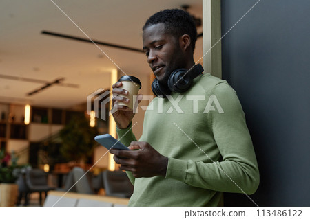 Minimal side view portrait of Black young man using smartphone leaning on wall in office lounge and drinking coffee copy space 113486122