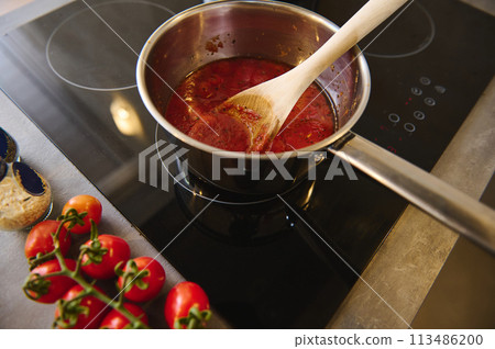 View from above wooden spoon inside a steel pan with boiling homemade sauces of ripe organic juicy red tomatoes. branch of fresh red tomato cherry on the kitchen counter near a black induction cooker View from above wooden spoon inside a steel pan with boiling homemade sauces of ripe organic juicy red tomatoes. branch of fresh red tomato cherry on the kitchen counter near a black induction cooker 113486200