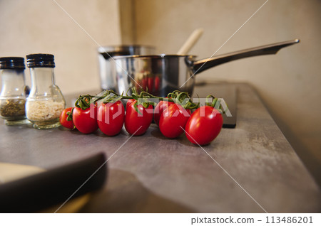 Still life with a branch of fresh ripe organic tomato cherry against blurred background of steel saucepan and bottles of condiments on the kitchen counter. Food background Still life with a branch of fresh ripe organic tomato cherry against blurred background of steel saucepan and bottles of condiments on the kitchen counter. Food background 113486201