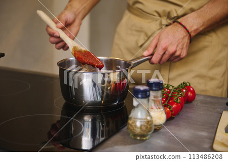 Close-up hands of a male chef holding a wooden spatula, stirring tomato juice with condiments, cooking passata according to traditional recipe in the home kitchen. Italian culinary. Mediterranean food 113486208