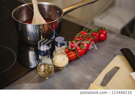 Two glass bottles of condiments, a branch of tomato cherry and steel pan with tomato sauce boiling and steaming on an electric stove in the home kitchen. Still life Two glass bottles of condiments, a branch of tomato cherry and steel pan with tomato sauce boiling and steaming on an electric stove in the home kitchen. Still life 113486210