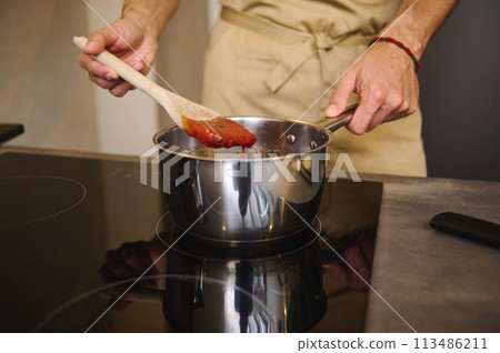 Close-up chef standing by electric induction stove, using wooden ladle, stirring boiling tomato juice, cooking homemade passata from organic juicy tomatoes in the kitchen 113486211