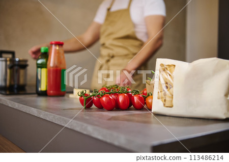 Selective focus on the bunch of red ripe organic tomato cherry and a paper packet with Italian pasta, against blurred male chef standing at induction stove, adjusting program while cooking dinner in 113486214