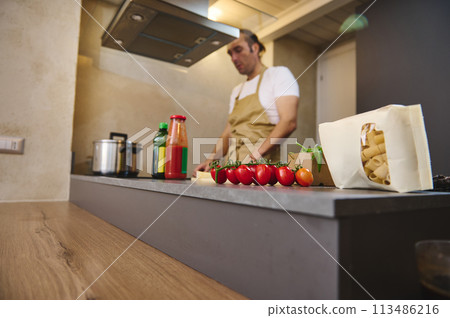 A bunch of red ripe organic tomato cherry and a packet with Italian pasta, against blurred male chef standing at induction stove, adjusting program while cooking dinner in modern home kitchen interior A bunch of red ripe organic tomato cherry and a packet with Italian pasta, against blurred male chef standing at induction stove, adjusting program while cooking dinner in modern home kitchen interior 113486216