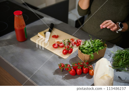 Close-up woman standing at kitchen counter with a fresh bunch of tomato cherry, arugula leaves and Italian pasta in paper packet. Healthy eating, culinary, diet concept. Copy advertising space 113486237