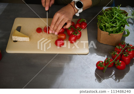 Overhead view of woman chopping tomato cherry on cutting board. A slice of delicious cheese, a bunch of organic tomatoes and fresh arugula leaves on the kitchen countertop. Italian Mediterranean food 113486240
