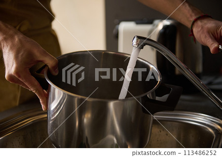 Man's hands filling a stainless steel saucepan with running water until it boils and cooks something. The concept of preparing food in the home kitchen Man's hands filling a stainless steel saucepan with running water until it boils and cooks something. The concept of preparing food in the home kitchen 113486252