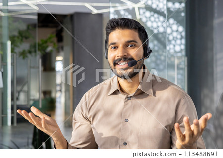 Cheerful Indian businessman wearing a headset communicating in a modern office setting. Engaged and professional, he looks directly at the camera. 113488691