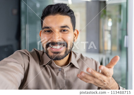 Confident businessman in a casual shirt smiling and talking on a phone while looking at the camera, standing in a modern office. 113488715