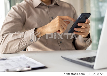 Midsection of a professional man using a smartphone while sitting at a modern office desk with a laptop. Focus on digital communication in a business setting. 113488726