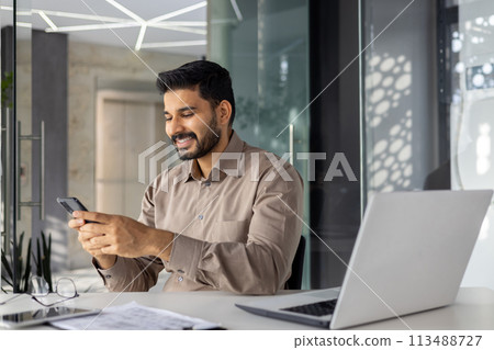 A cheerful young businessman engages with his smartphone, seated in a well-lit, contemporary office space, showcasing a blend of technology and casual corporate style. 113488727