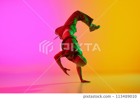 Dynamic photo of young man performing freestyle moves in neon light against gradient pink-yellow background. 113489010