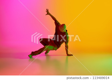 Young man, dressed streetwear performing freestyle moves in motion in neon light against gradient pink-yellow background. Young man, dressed streetwear performing freestyle moves in motion in neon light against gradient pink-yellow background. 113489014