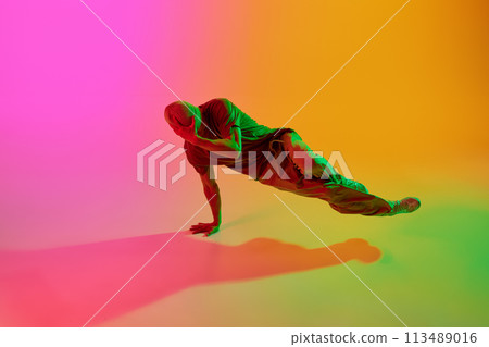 Young athlete man, dressed streetwear dancing freestyle moves in motion in neon light against gradient pink-yellow background. Young athlete man, dressed streetwear dancing freestyle moves in motion in neon light against gradient pink-yellow background. 113489016
