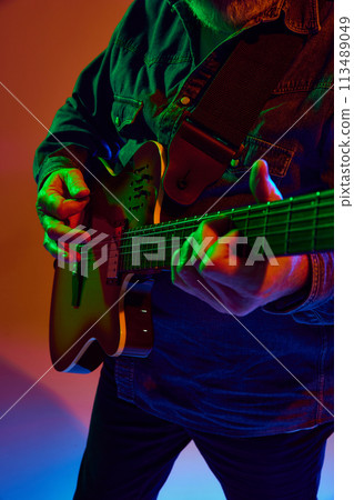 Cropped photo of elderly man guitarist performing chords of rock-ballad in colorful neon light against orange studio background. 113489049