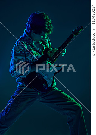 Young talented guitarist in retro attire performing in green blue stage neon light against gradient studio background. 113489234