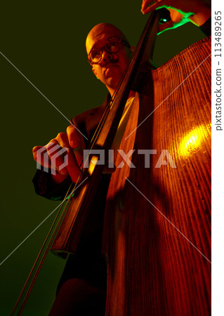 Artistic, talented young bass player performing jazz composition in red-green neon light against gradient studio background. Artistic, talented young bass player performing jazz composition in red-green neon light against gradient studio background. 113489265