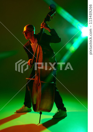 Double bass player performing contemporary jazz composition in red-green neon light against gradient studio background. Double bass player performing contemporary jazz composition in red-green neon light against gradient studio background. 113489266