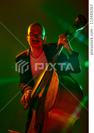Stylish, contemporary bassist in glasses performs in red-green neon stage light against gradient studio background. Stylish, contemporary bassist in glasses performs in red-green neon stage light against gradient studio background. 113489267