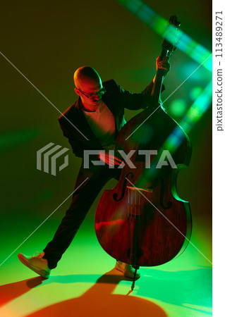 Talented cellist performing jazz compositions in red-green neon light against gradient studio background. Talented cellist performing jazz compositions in red-green neon light against gradient studio background. 113489271