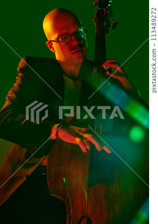 Bald man, talented cellist performing jazz compositions in red-green neon light against gradient studio background. Bald man, talented cellist performing jazz compositions in red-green neon light against gradient studio background. 113489272