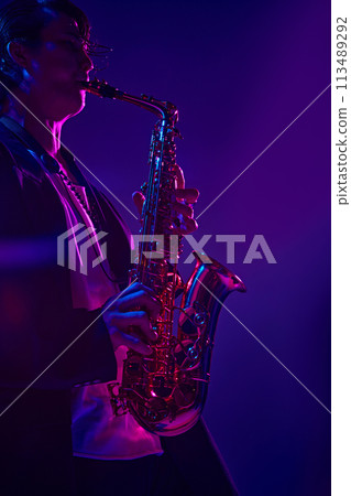 Portrait of young attractive man in dark suit playing saxophone in vibrant pink neon light against dark studio background. Portrait of young attractive man in dark suit playing saxophone in vibrant pink neon light against dark studio background. 113489292