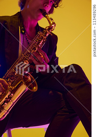 Cropped portrait of solo player in black suit playing sax in neon light against vibrant yellow studio background. Cropped portrait of solo player in black suit playing sax in neon light against vibrant yellow studio background. 113489296