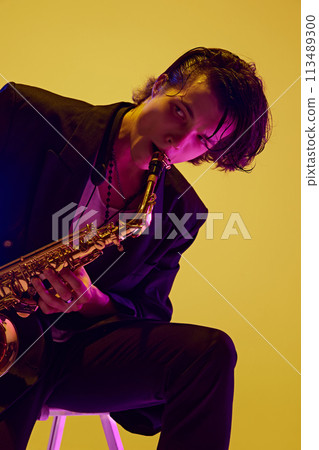 Portrait of young attractive man in dark suit playing saxophone in neon light against vibrant yellow studio background. Portrait of young attractive man in dark suit playing saxophone in neon light against vibrant yellow studio background. 113489300