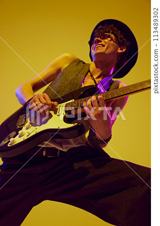 View from below of young talented man playing electric guitar in neon light against vibrant yellow studio background. 113489302