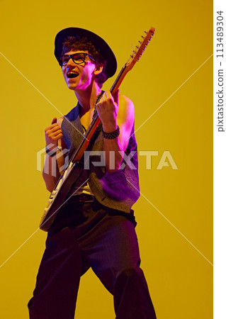 Overjoyed young man in retro hat and glasses playing with drive rock-n-roll music in neon light against yellow studio background. 113489304
