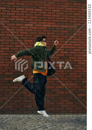 Trendy dressed young guy in sporting jacket and sunglasses jumping in motion against brick backdrop. Trendy dressed young guy in sporting jacket and sunglasses jumping in motion against brick backdrop. 113489310