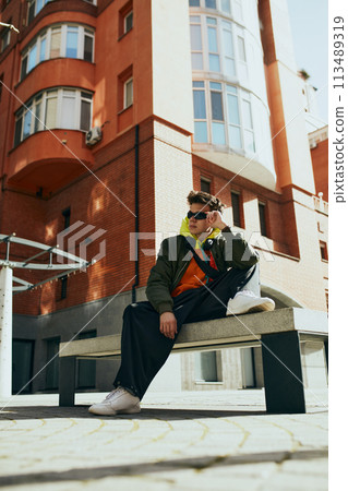 Creative portrait of young attractive man dressed oversize clothes, stylish accessories against cityscape background. 113489319