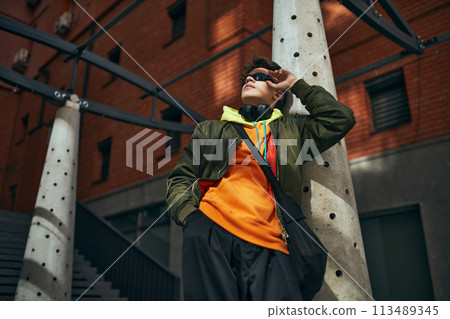 View from below young cool attitude man posing wearing vibrant clothe and stylish glasses posing against urban background. 113489345