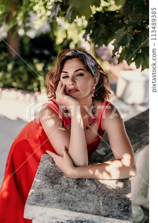 A pretty woman in a red silk dress and a bandage on her head smiles against the background of the leaves of a tree. She is leaning on the coop and looking into the camera. Vertical photo. 113489385