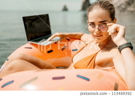 Woman laptop sea. Freelancer woman in sunglases floating on an inflatable big pink donut with a laptop in the sea. People summer vacation rest lifestyle concept 113489392