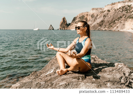 Yoga on the beach. A happy woman meditating in a yoga pose on the beach, surrounded by the ocean and rock mountains, promoting a healthy lifestyle outdoors in nature, and inspiring fitness concept. Yoga on the beach. A happy woman meditating in a yoga pose on the beach, surrounded by the ocean and rock mountains, promoting a healthy lifestyle outdoors in nature, and inspiring fitness concept. 113489433