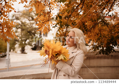 A blonde woman is holding a bunch of yellow leaves in her hand. She is standing in front of a tree with leaves that are changing colors. The scene has a peaceful and serene mood. 113489444