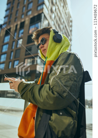 Young man in trendy sunglasses and headphones and bright casual attire stands confidently outside and using phone. 113489672