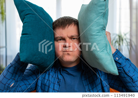 Irritated Caucasian man covers ears with cushions annoyed by noisy loud music neighbors repair work Irritated Caucasian man covers ears with cushions annoyed by noisy loud music neighbors repair work 113491940