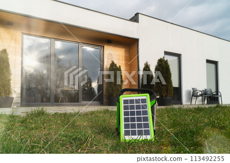 Small solar panel in green frame on lawn, contemporary home with large windows in background 113492255