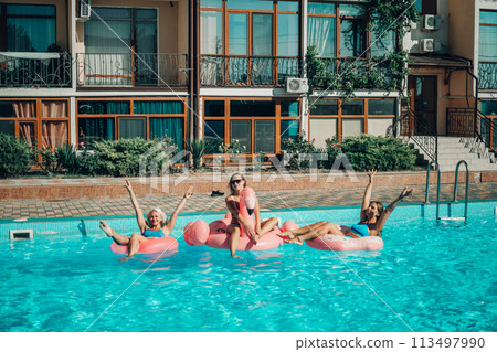 A group of people are in a pool with pink and white floats. Scene is fun and lighthearted. 113497990