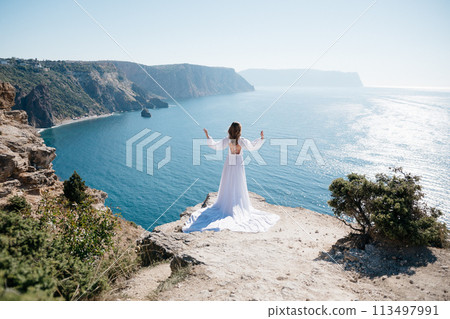 A woman stands on a cliff overlooking the ocean in a white dress. She is looking out at the water and she is in a state of peace and serenity. Concept of calm and tranquility. 113497991
