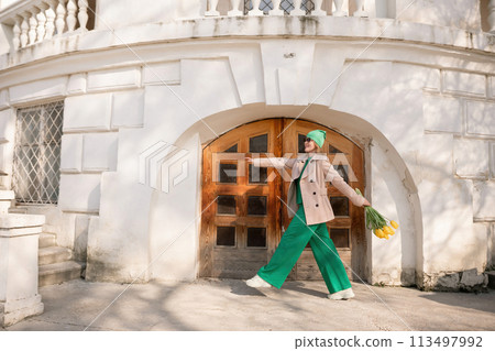 A woman in a green suit and green pants is walking down a street with a bouquet of yellow flowers in her hand. 113497992