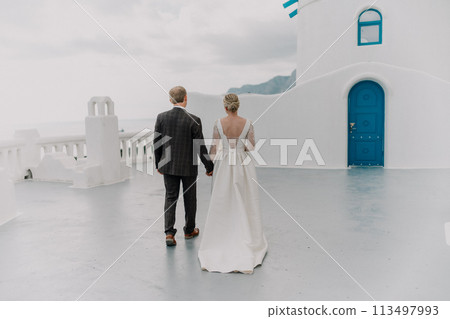 A bride and groom are walking down a path in front of a blue door. The bride is wearing a white dress and the groom is wearing a suit. Scene is romantic and happy. A bride and groom are walking down a path in front of a blue door. The bride is wearing a white dress and the groom is wearing a suit. Scene is romantic and happy. 113497993