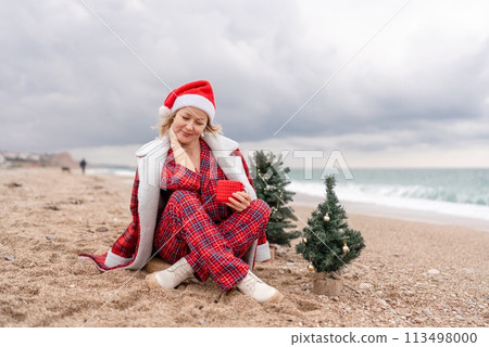 Sea Lady in Santa hat plaid shirt with a red mug in her hands enjoys beach with Christmas tree. Coastal area. Christmas, New Year holidays concep Sea Lady in Santa hat plaid shirt with a red mug in her hands enjoys beach with Christmas tree. Coastal area. Christmas, New Year holidays concep 113498000