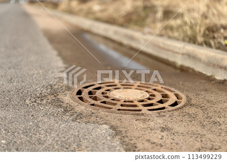 A old iron hatch with a pattern above an observation well is located on auto road in daytime. Close-up 113499229