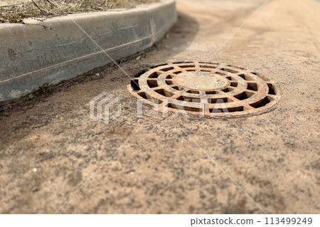 A rusty iron hatch with a pattern above an observation well is located on auto road in daytime. Close-up 113499249