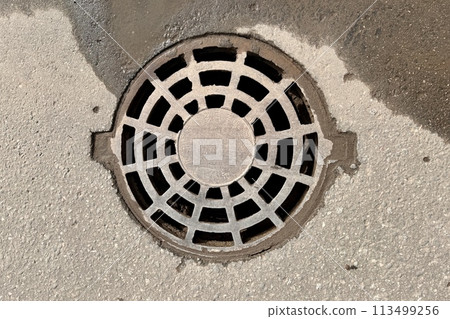 A iron hatch with a pattern above an observation well is located on auto road in daytime. Close-up 113499256