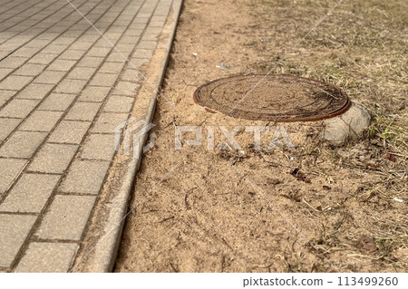 A iron manhole above an observation well is located next to a dry grass pedestrian road during the daytime. Close-up 113499260